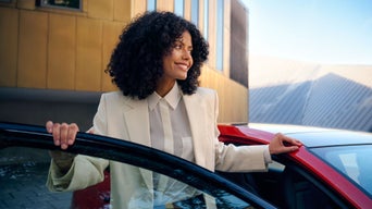 Frau mit lockigem Haar steigt lächelnd aus einem roten Auto vor einem modernen Gebäude mit goldfarbener Fassade.