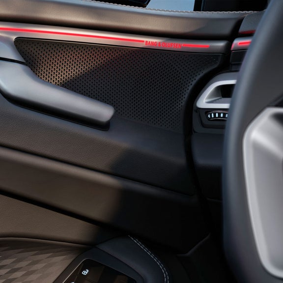 Close-up of a car door interior featuring a Bang & Olufsen speaker, sleek black leather, and illuminated red accents.
