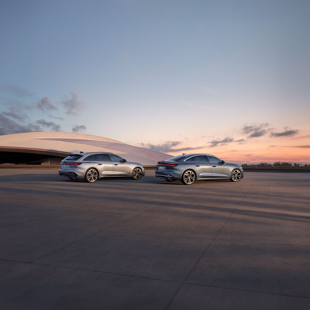 Silberner Audi A5 Avant und blaue A5 Limousine vor moderner Architektur bei Sonnenuntergang. 