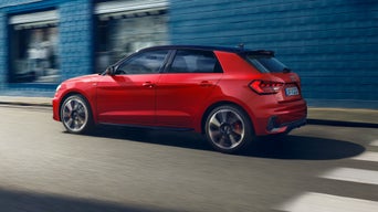 Une Audi A1 Sportback rouge est montrée de trois quarts arrière, roulant dans une rue, avec un bâtiment flou en arrière-plan suggérant le mouvement.