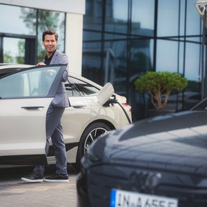 A person in a suit smiles while opening a car door in a modern setting. A black car in the foreground, with glass building reflections and greenery.