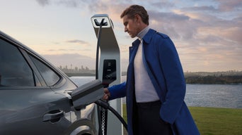 A person in a blue coat connects a charging cable to an electric vehicle at a modern charging station, with a city skyline in the background.