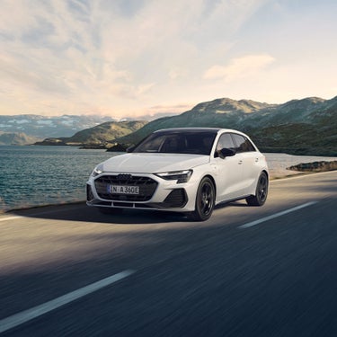 Weißer Audi A3 Sportback TFSI e fährt auf einer Straße entlang eines Sees mit Bergen im Hintergrund.