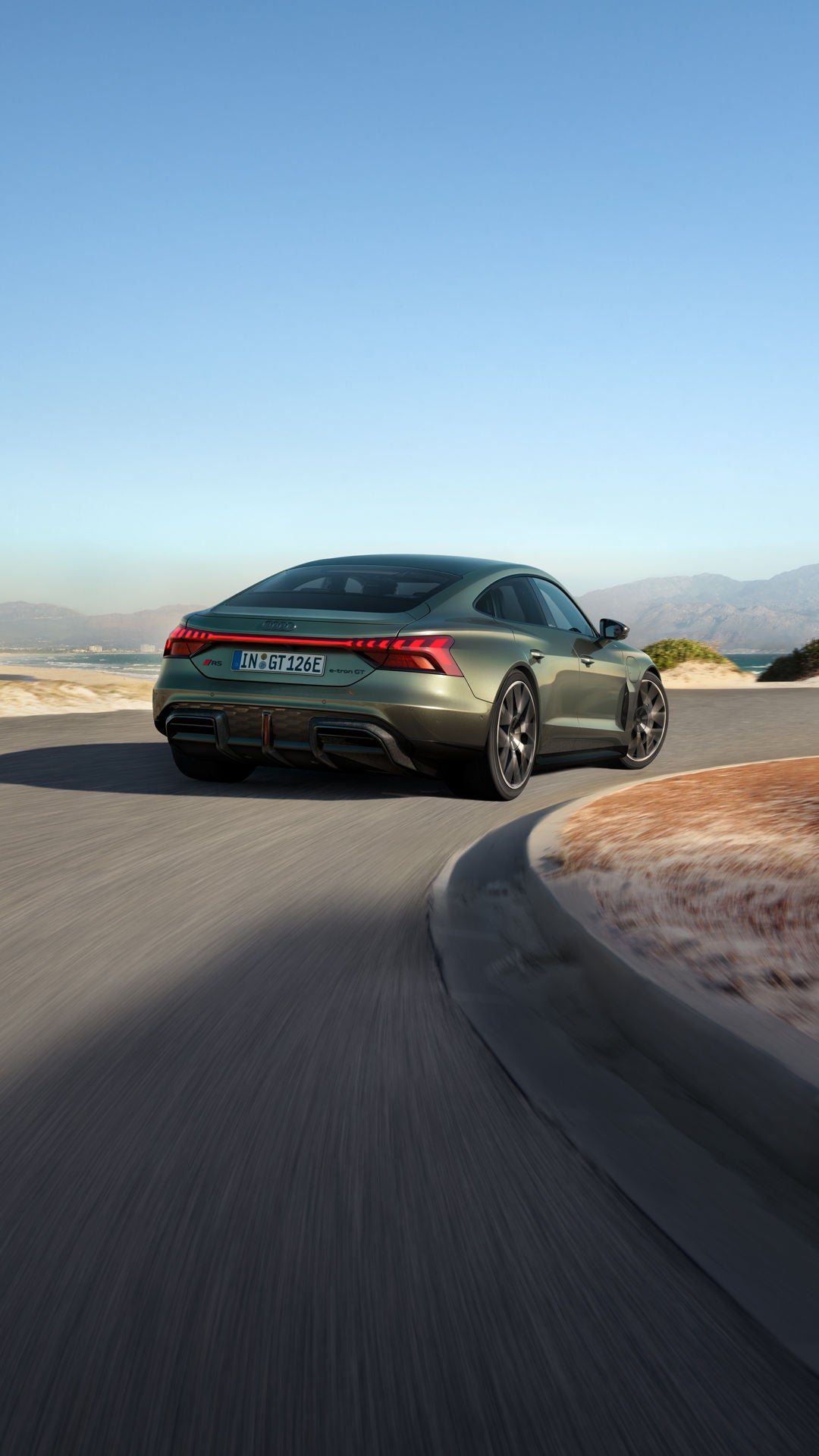 Um Audi RS e-tron GT performance verde percorre uma estrada costeira sinuosa, com montanhas, o mar e um céu limpo ao fundo.