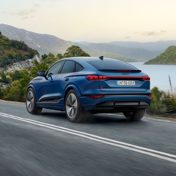 Schräge Heckaufnahme des SQ6 Sportback e-tron, welcher eine Straße an einem See in einer bergigen Landschaft entlangfährt.