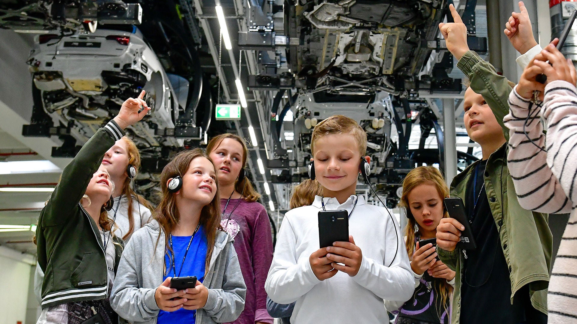 A group of children wearing headsets is standing beneath production equipment and looking up; one boy is looking at a smartphone.
