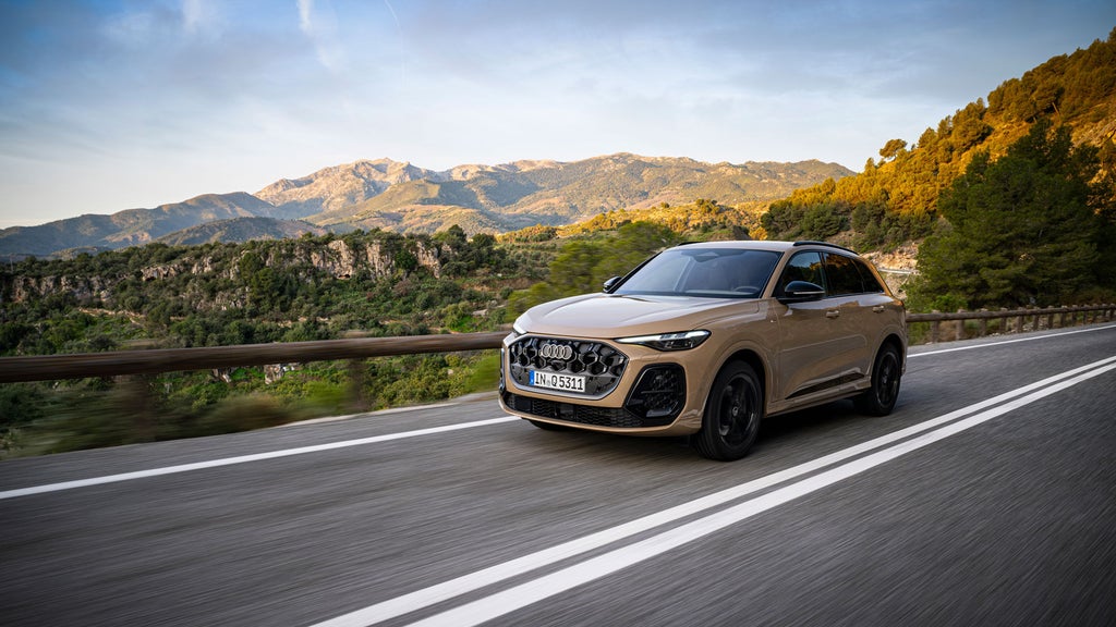 Ein Audi Q5 SUV fährt eine Bergstraße entlang. Im Hintergrund grün bewachsene Hügel in goldfarbenem Sonnenlicht.