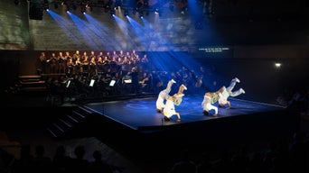 Vier Tänzer in weißer Kleidung bewegen sich auf der Bühne vor Orchester und Chor, blaues Bühnenlicht.