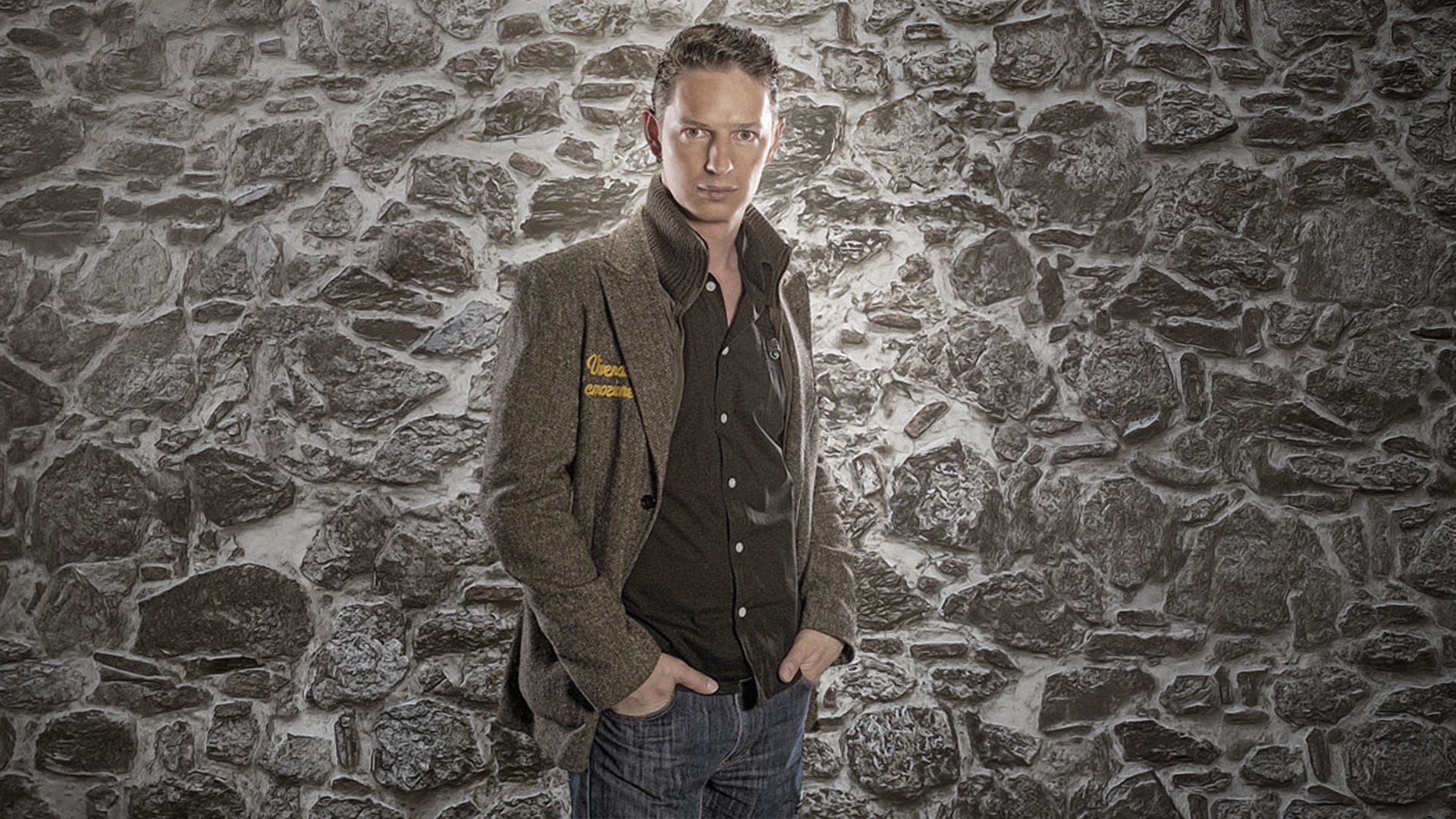 Mann in Jacke und Jeans steht mit den Händen in den Taschen vor einer grauen Steinwand.