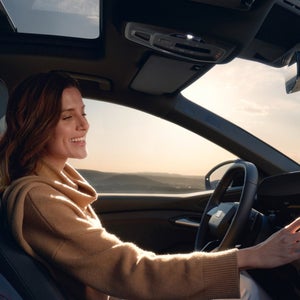 A woman in a camel sweater looks relaxed and happy while driving a car at sunset. The interior is sleek, with a clear view through the windshield.