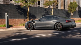 Ein Audi RS e-tron GT lädt an einer Straßenladestation vor einer modernen Fassade mit Bäumen und Schatten.