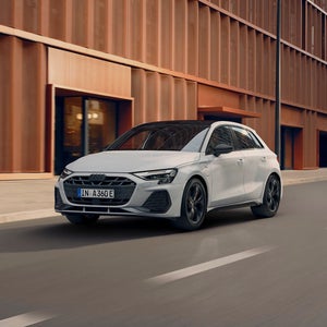 A white Audi A3 Sportback TFSI e drives along a modern urban street, set against a stylish brown building facade.