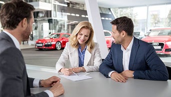 Three people at a table in a car dealership, with a woman signing a document; red Audi cars in the background. 