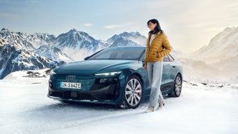 Frau in gelber Jacke steht neben blauem Audi auf verschneiter Fläche vor Bergkulisse.