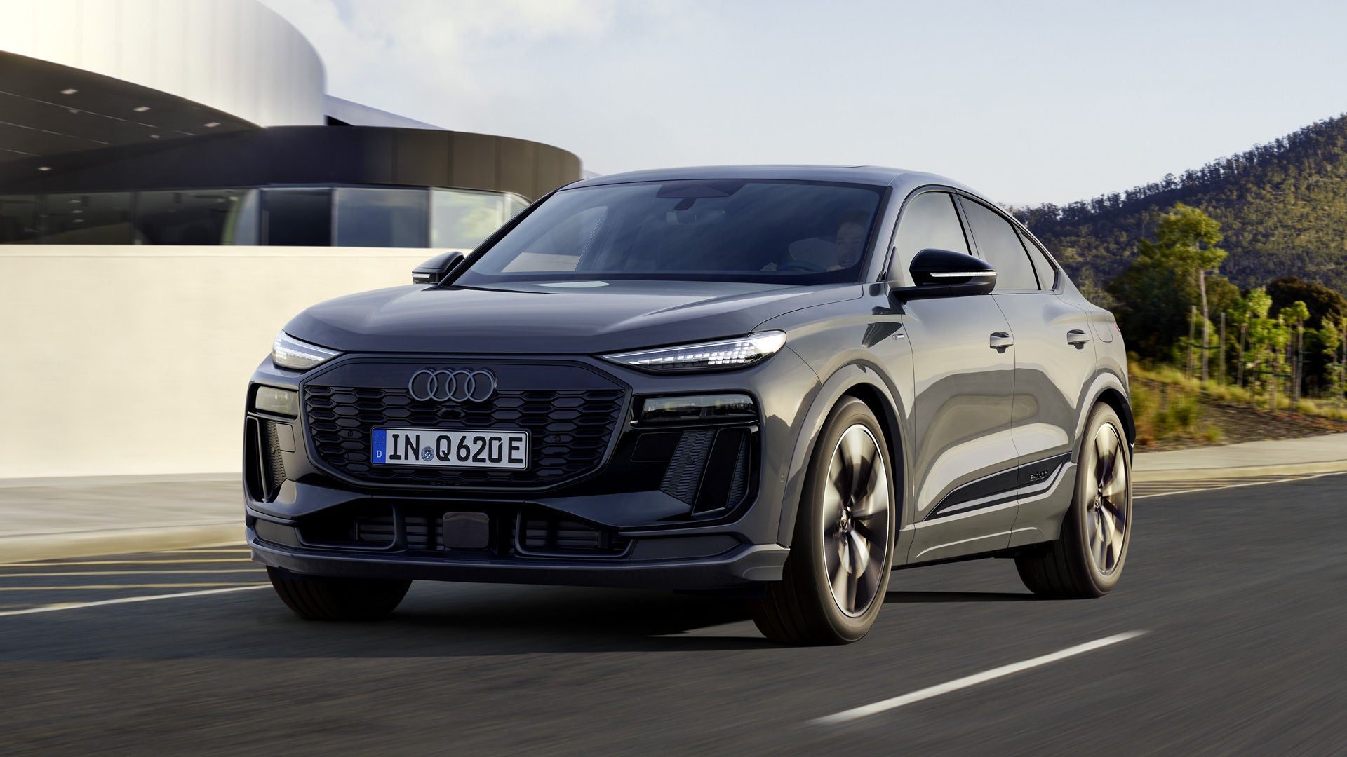 Schräges Frontprofil des Q6 Sportback e-tron, welcher auf einer Straße neben einem modernen Haus fährt.