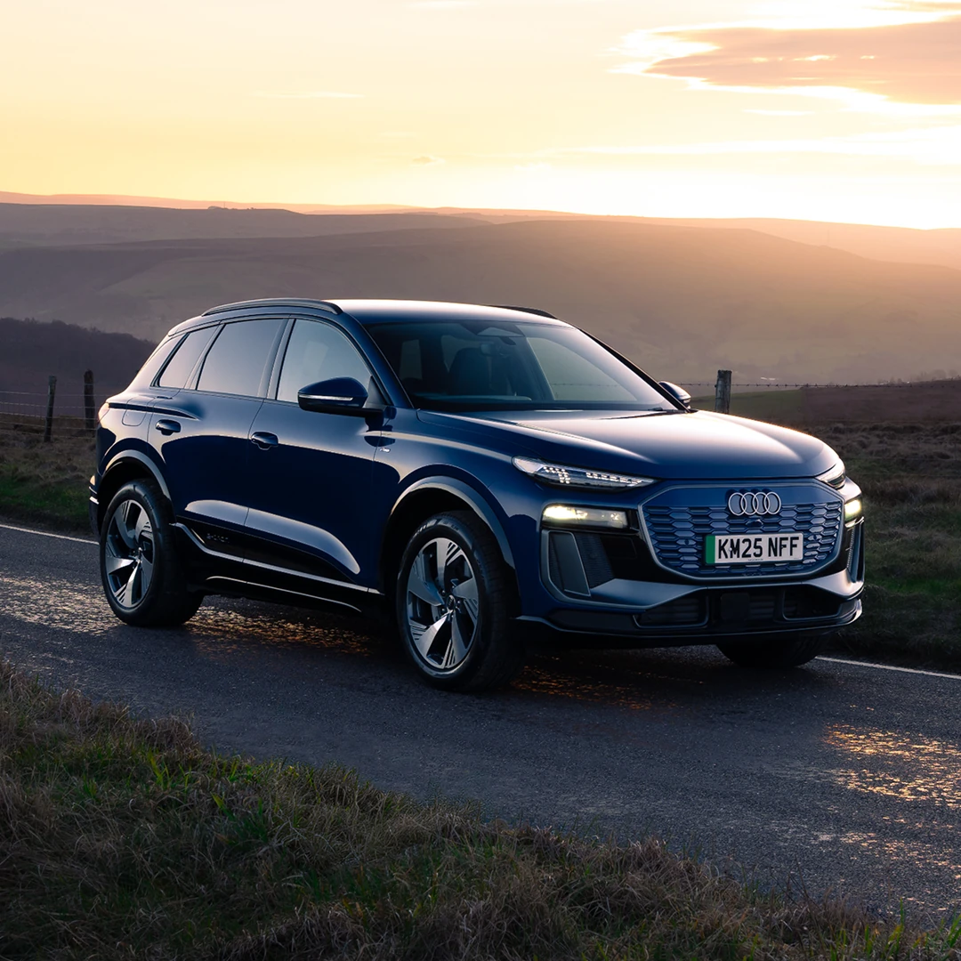 A dark blue Audi SUV drives on a coastal highway at sunset. The road curves gently with the ocean to the right and cliffs to the left. The scene is serene and dynamic.