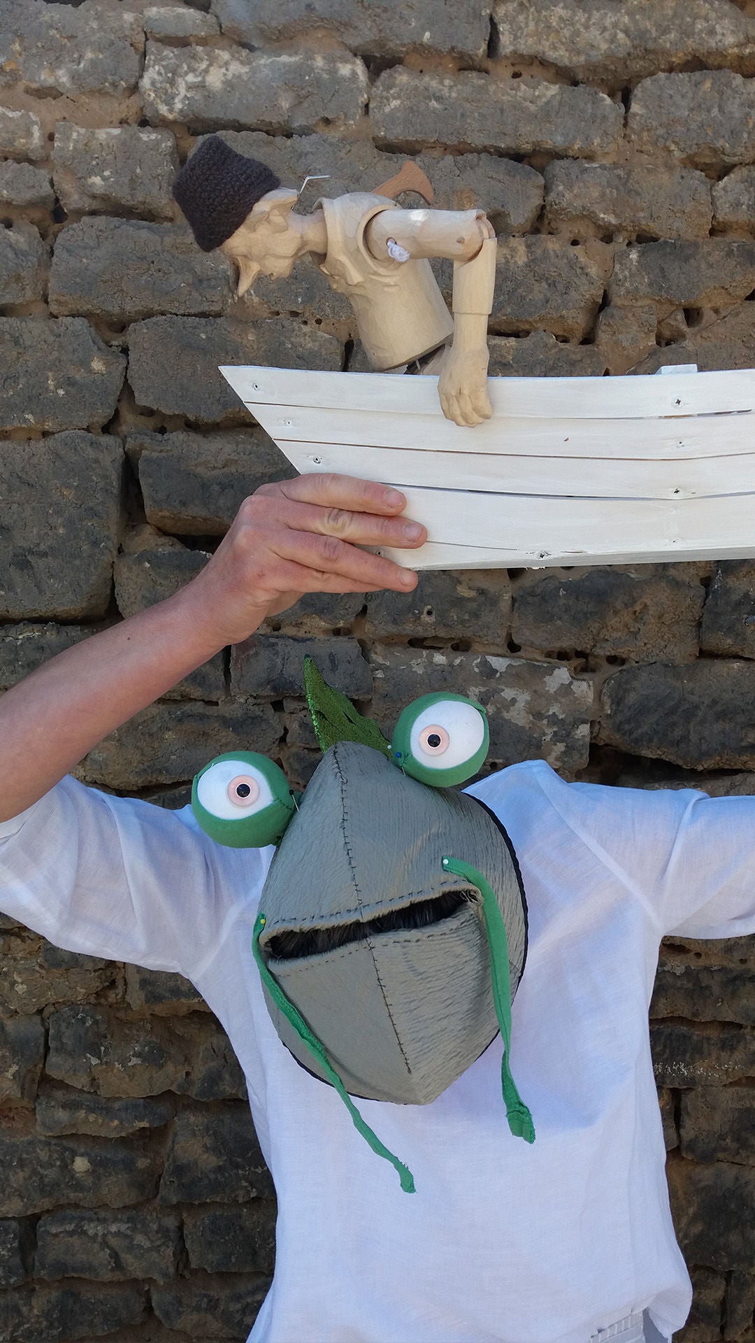 Person mit Fischmaske hält ein kleines weißes Boot mit einer Figur über dem Kopf vor einer Steinmauer.