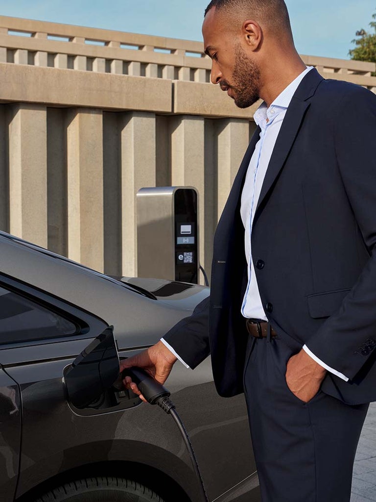 Farbige Person im Anzug steckt Ladekabel an grauen Audi A8 L 60 TFSI e an, Ladesäule und beige Betonwand im Hintergrund.