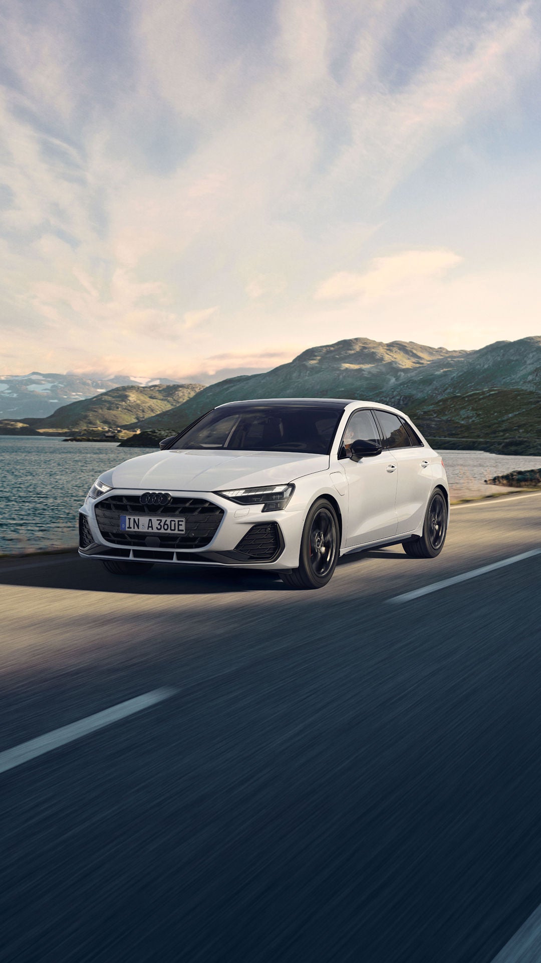 A white Audi car drives along a scenic lakeside road, flanked by mountainous terrain under a clear sky. The setting conveys freedom and adventure.