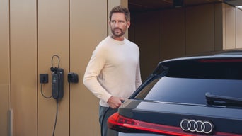 Der S6 Avant e-tron steht vor einem modernen Haus. An der Hausfassade ist eine Ladestation montiert, welche von einem Mann mittleren Alters an das Fahrzeug angeschlossen wird.