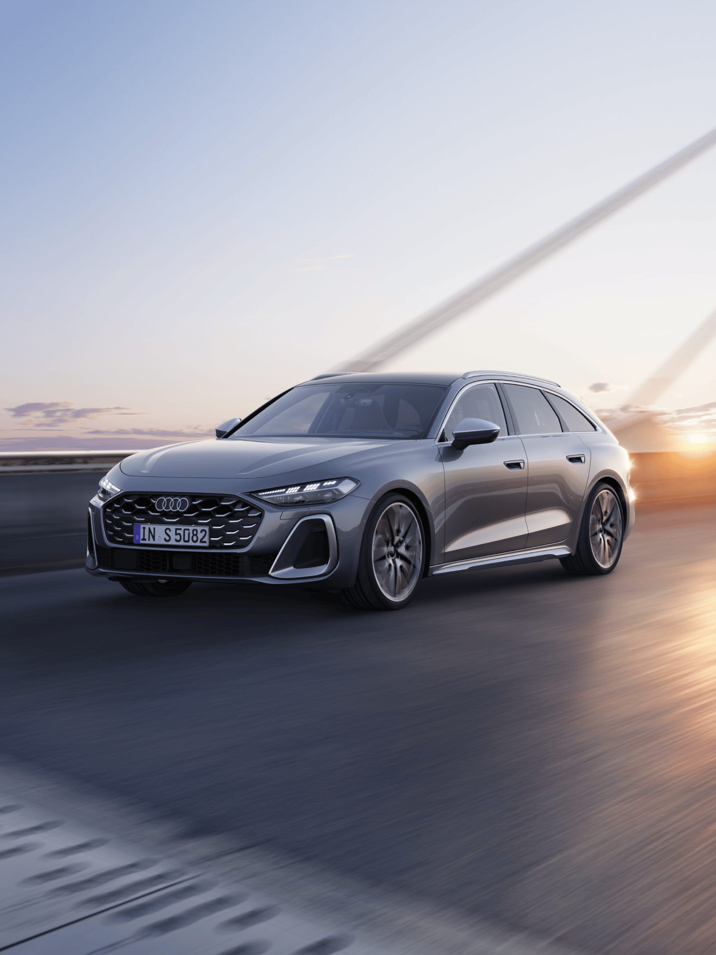 A silver Audi S5 Avant car driving on a road at dusk, with sun setting in the background.
