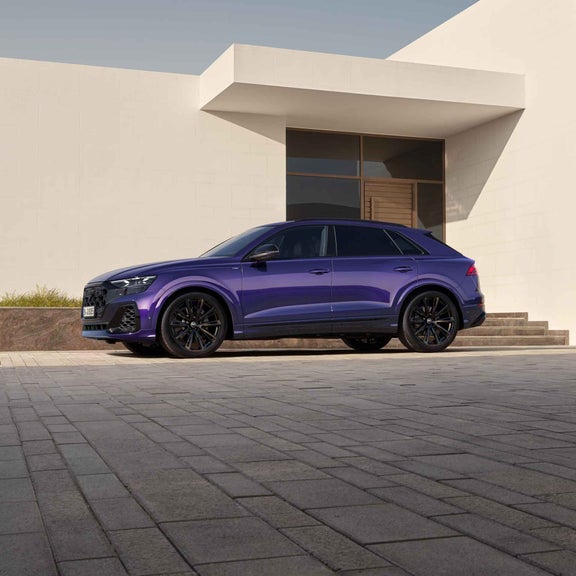 Der velvet-lila Q8 SUV parkt auf gepflasterten Steinen vor einem modernen Haus. Im Hintergrund ist die Innenstadt bei Tag zu sehen.