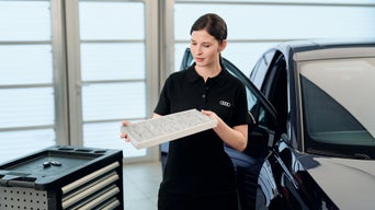 Eine Audi Mitarbeiterin hält einen Fahrzeugfilter neben einem Audi Fahrzeug in der Werkstatt