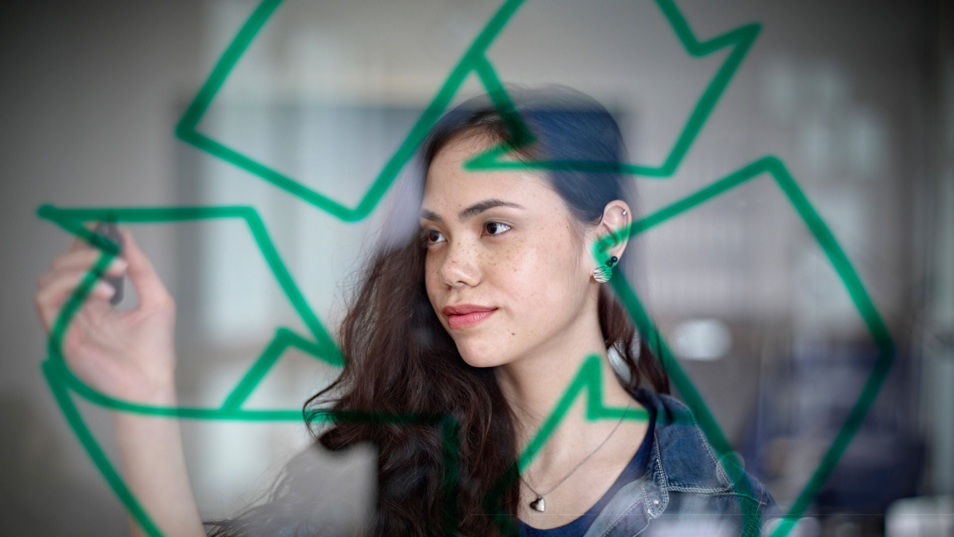 Eine junge Frau malt das Recycling-Symbol aus dreieckig angeordneten, gebogenen Pfeilen an eine durchsichtige Tafel