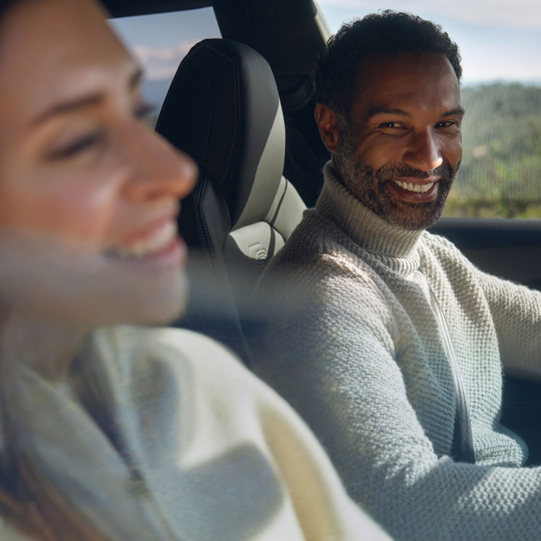 A person wearing a light-colored sweater is driving a car, focused on the road, with another passenger visible in the foreground.