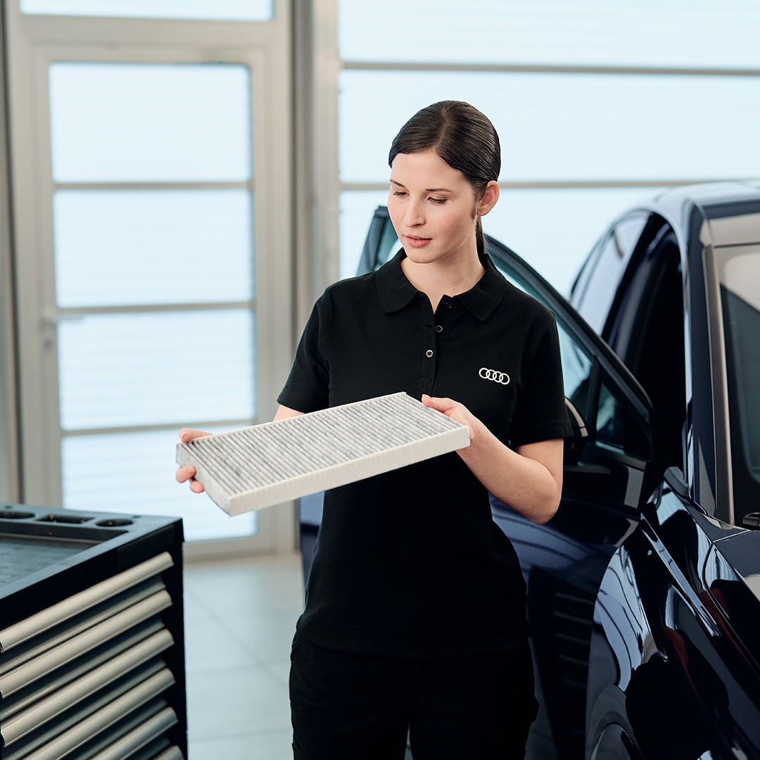 Audi-Technikerin betrachtet rechteckigen Innenraumfilter neben Fahrzeug in heller Werkstatt.
