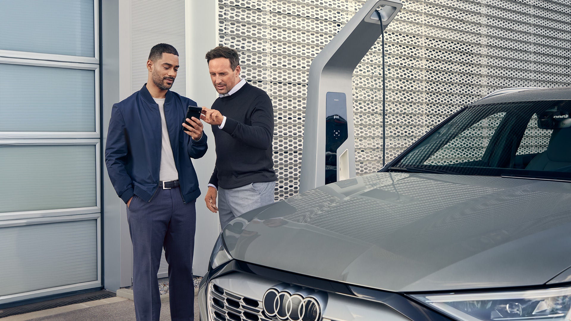 Ein silbernes Audi Modell läd an einer Elektrosäule und ein Verkaufsberater zeigt einem anderen Mann im Anzug auf dem Smartphone etwas
