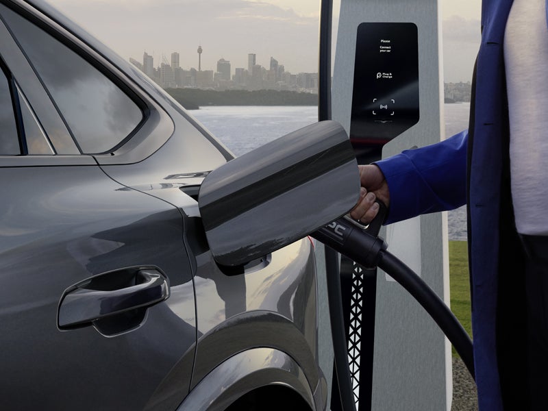 Nahaufnahme der Ladebuchse des Q6 Sportback e-tron, in welche eine Frau gerade ein Ladekabel einsteckt. Im Hintergrund befindet sich das Meer, sowie die Skyline einer Stadt.