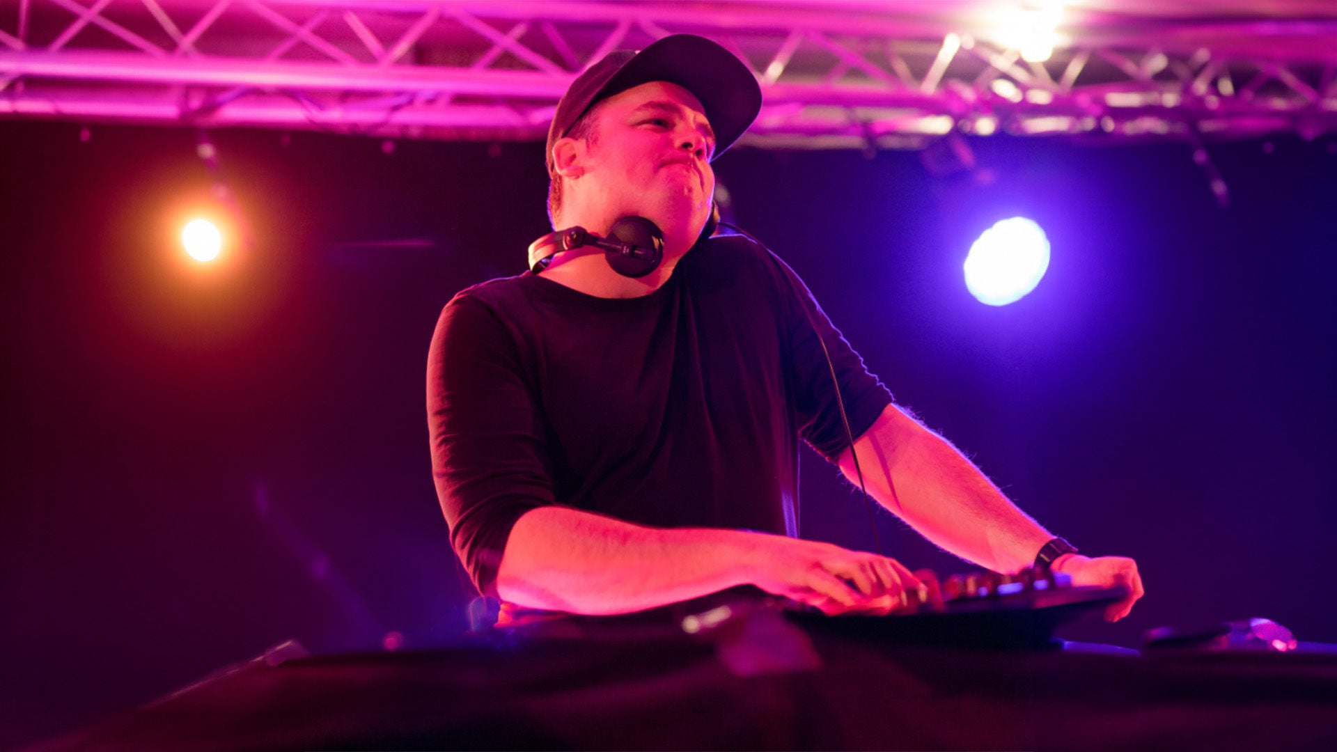 DJ mit Cap und Kopfhörern am Hals steht an einem Mischpult, beleuchtet von pinken und violetten Bühnenlichtern.