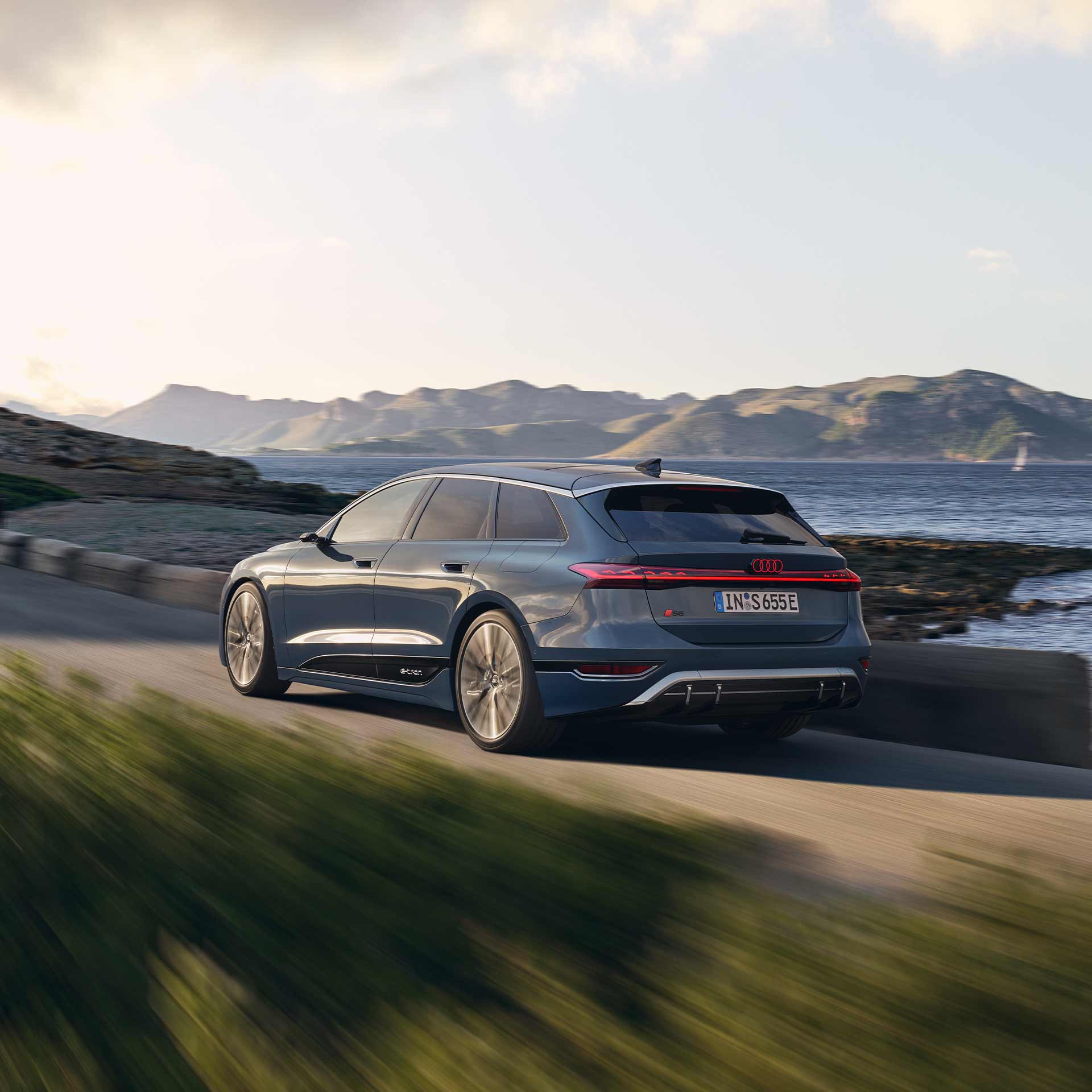 Dunkelblauer Audi S6 Avant e-tron fährt an einer Küstenstraße entlang, im Hintergrund Berge und Meer.