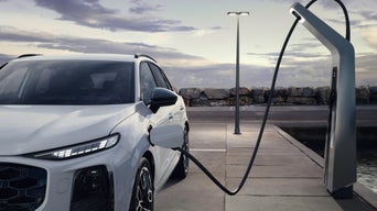 Woman in orange dress and white jacket walking beside a silver Audi RS e-tron GT car on a coastal road.