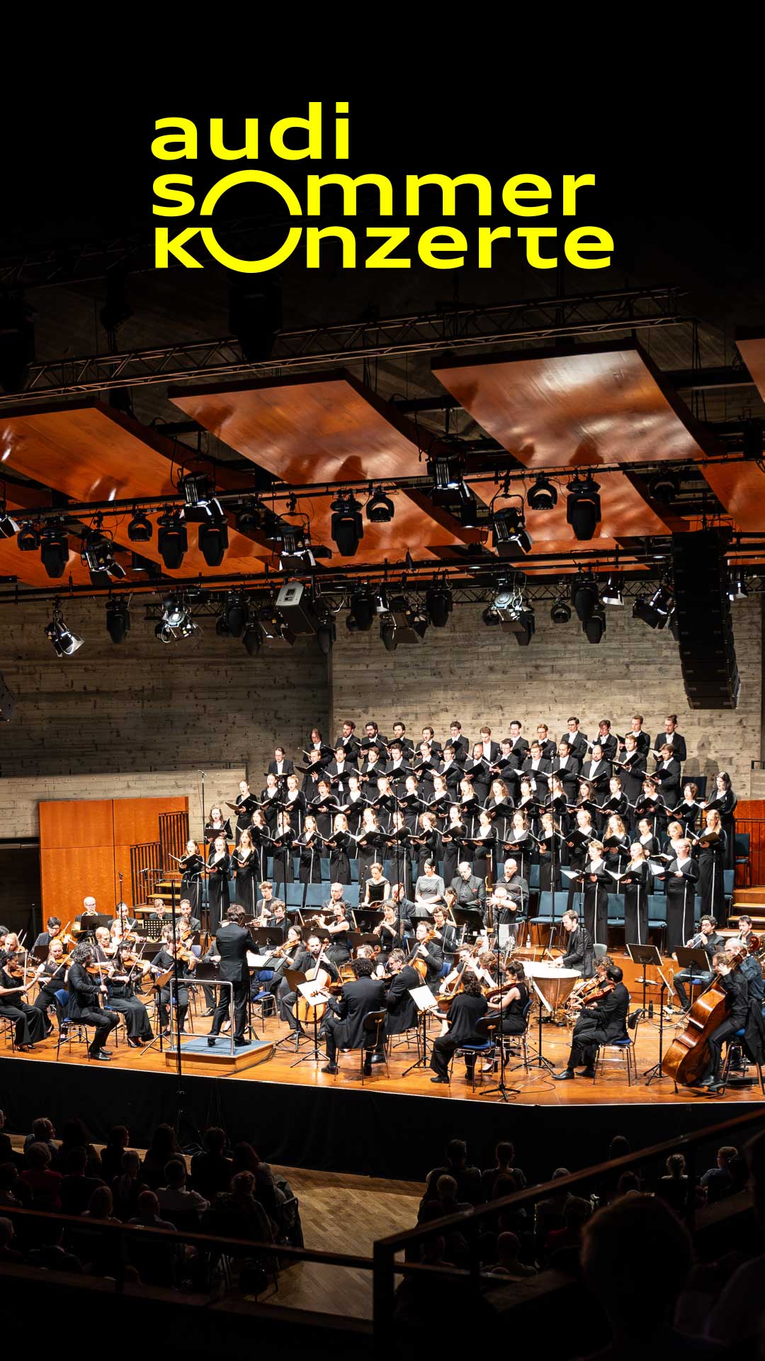 Konzertsaal mit Orchester und Chor auf der Bühne, Publikum im Vordergrund, Text „audi sommerkonzerte“ eingeblendet.