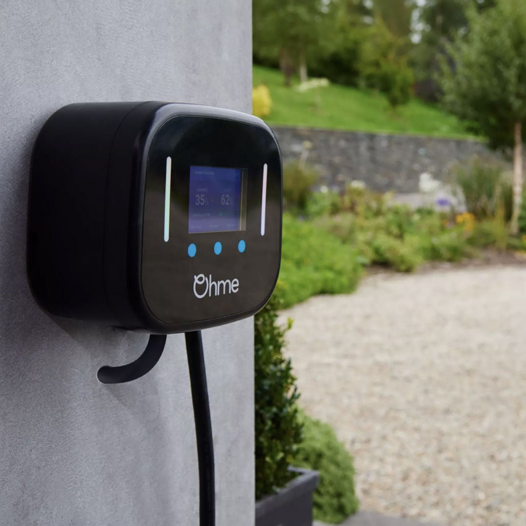 An Ohme electric vehicle charger mounted on a textured wall, surrounded by a lush garden and gravel walkway.