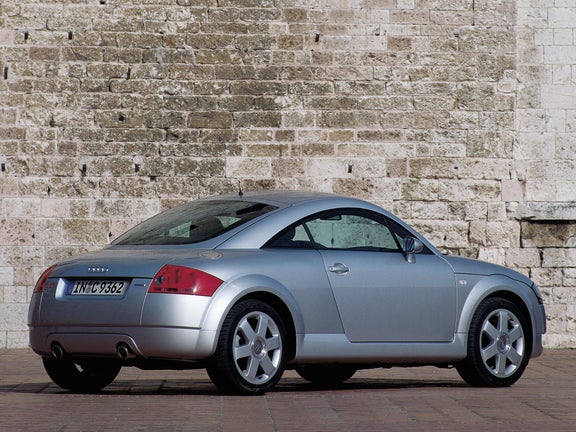 A rear side view of a grey Audi TT in front of a stone wall