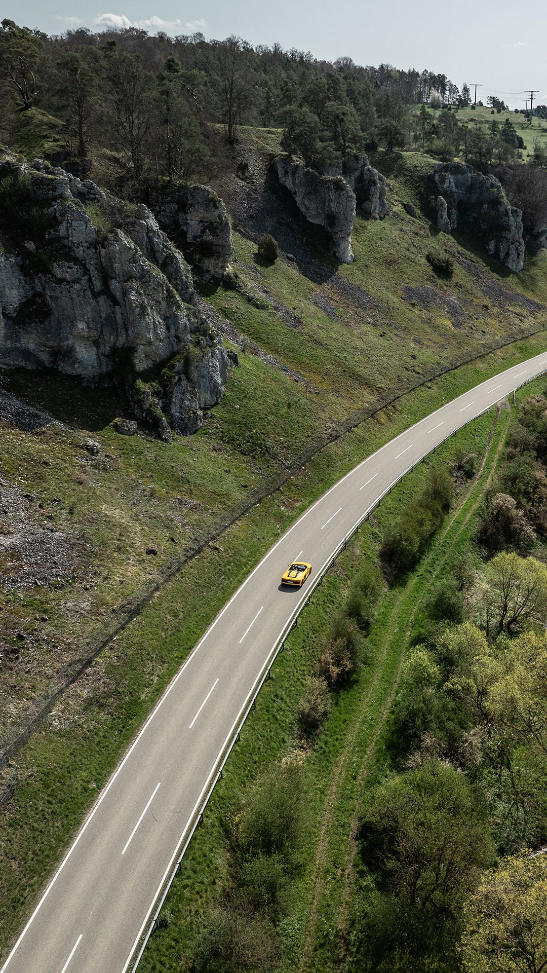 Aerial view of a yellow sports car on a winding road between rocks and the riverbank.