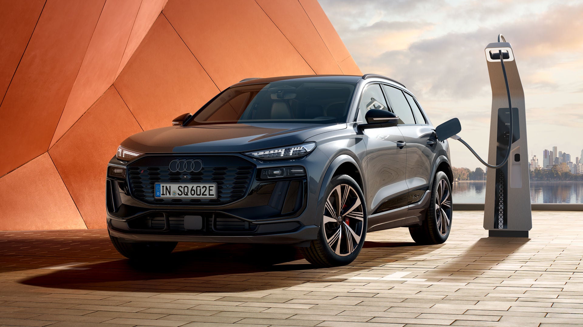 Der SQ6 SUV e-tron steht vor einer orangenen Gebäudefassade und ist dabei an eine Ladestation angeschlossen. An der Seite des Fahrzeugs lehnt sich eine junge Frau an und schaut auf ihr Handy.&nbsp;