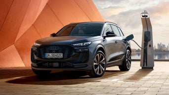 Audi SQ6 e-tron, widziane z przodu pod kątem, stoi przy pomarańczowej fasadzie budynku, podłączone do stacji ładowania. W tle widoczne są morze i panorama miasta.
