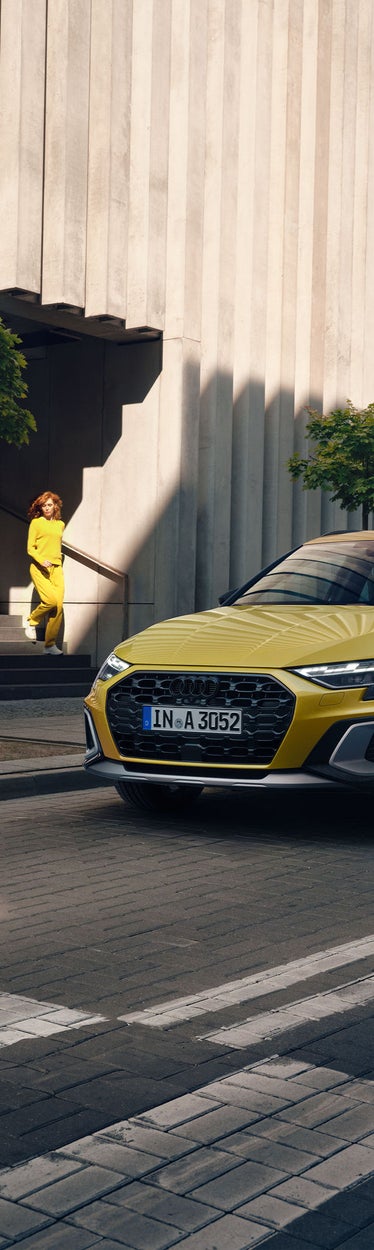Gelber Audi A3 allstreet steht am Straßenrand, eine Frau in gelber Kleidung geht eine Treppe im Hintergrund hinab.