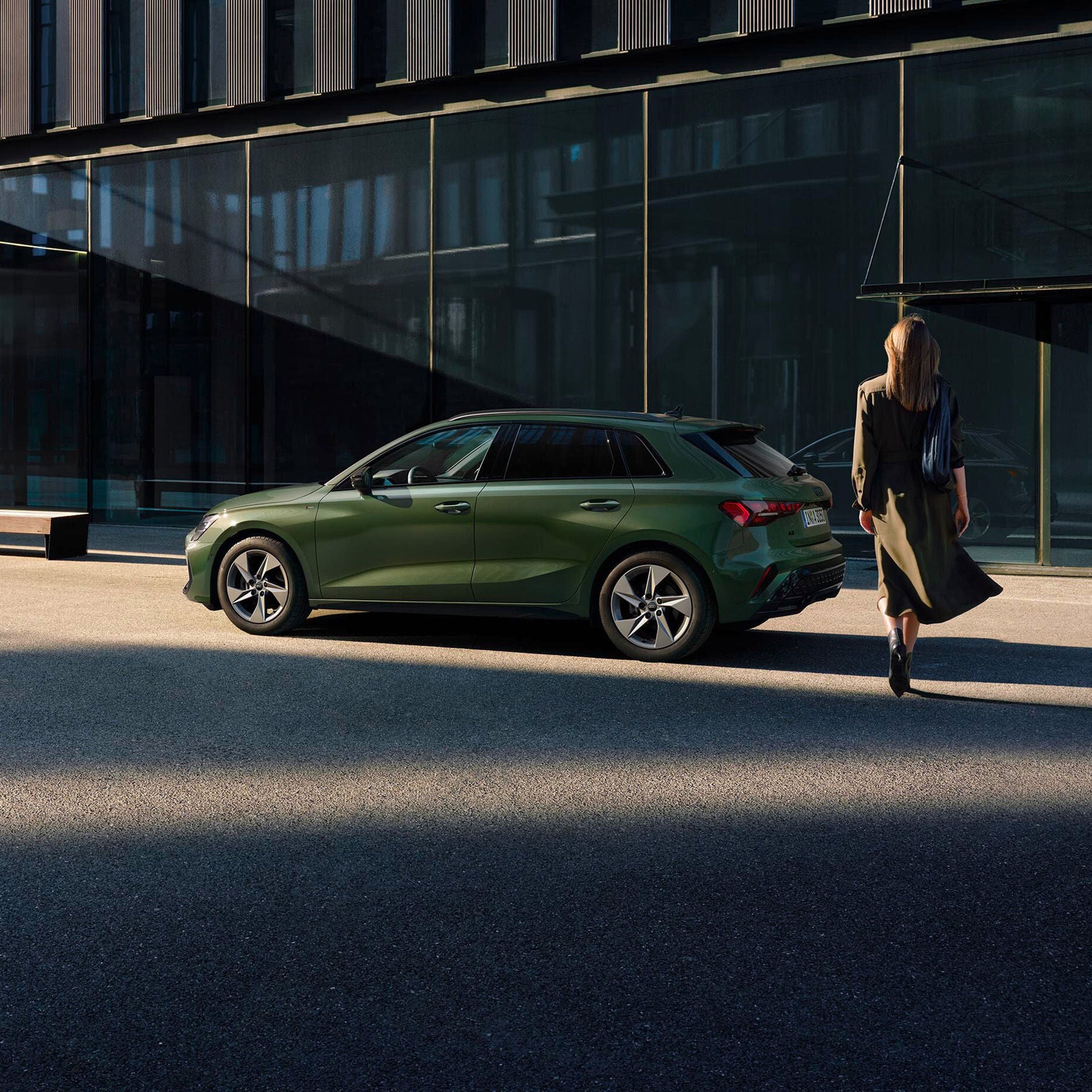 Eine Frau geht auf ihren grünen Audi A3 zu, der vor einem Gebäude mit Glasfassade in der Abendsonne steht.