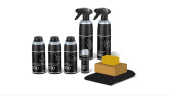 Verschiedene Sprays und Pflegemittelflaschen in schwarz mit hellem Audi Logo, rechts daneben zwei gelbe Schwämme und eine schwarze Tasche.