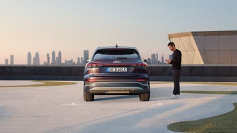 Ein Mann schaut auf sein Handy und steht neben einem geparkten Audi Q4 e-tron auf einem Dachparkplatz, im Hintergrund eine Skyline bei Sonnenuntergang.