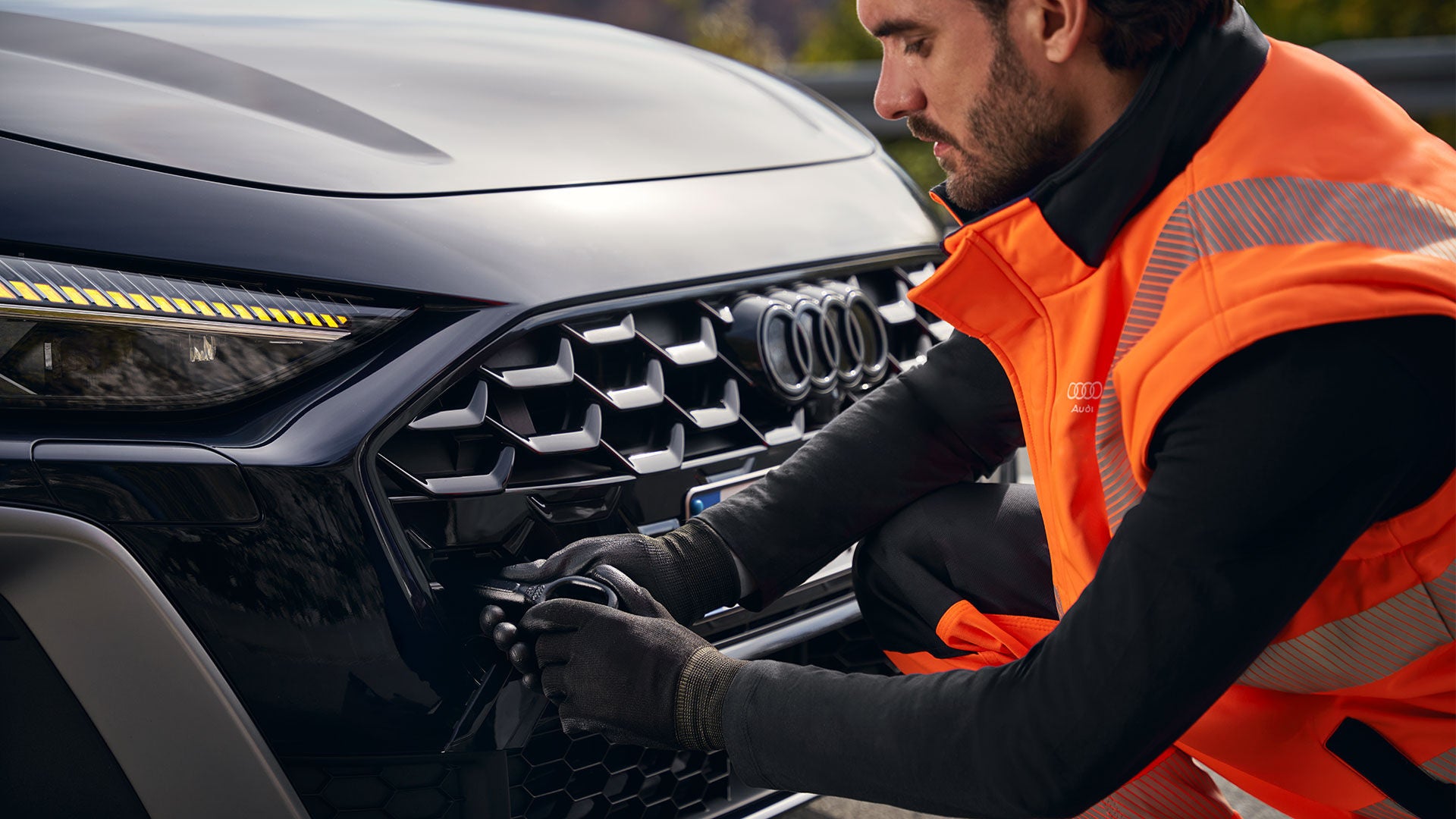 Ein Mann in orangener Warnweste kniet an der Motorhaube eines Audi und schraubt den Abschlepphaken in die dafür vorgesehene Öse.