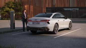 Das Bild zeigt einen weißen Audi A6 Limousine e-hybrid, der an eine Ladestation angeschlossen ist und den Einstieg der Marke in den Markt für Elektrofahrzeuge unterstreicht. Eine Frau im Geschäftsanzug steht in der Nähe des Autos und der Ladestation.