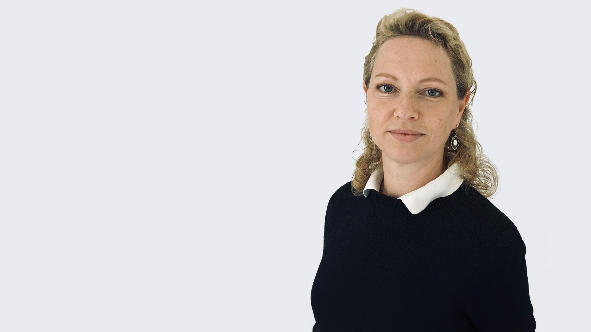 Iris Sasirmaz: Porträt einer Frau mit blondem, zurückgebundenem Haar, schwarzem Oberteil mit weißem Kragen, neutraler Blick vor hellem Hintergrund.