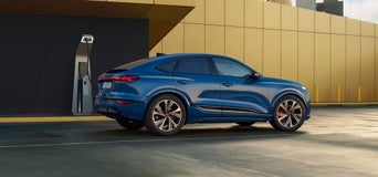 Der SQ6 Sportback e-tron steht seitlich auf einem Parkplatz vor einer gelben Fassade und ist dabei an eine Ladestation angeschlossen.