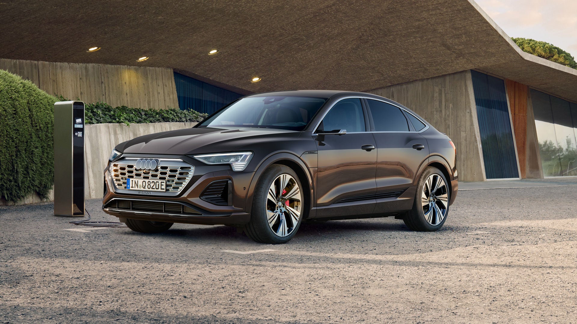 Ein brauner Audi Q8 e-tron steht an einer Ladestation vor einem modernen Gebäude mit schrägem Dach und großer Glasfassade.